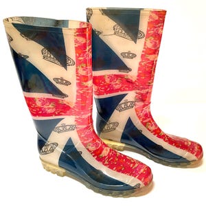 May include: A pair of mid-calf rain boots featuring a Union Jack flag pattern combined with a pink floral print and black crown illustrations. The boots have a glossy finish, a rounded toe, and a clear, textured rubber sole for traction.