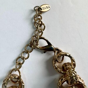 Puede incluir: Primer plano de un collar dorado con cierre de mosquet&oacute;n y eslabones de cadena. El collar presenta un dise&ntilde;o texturizado y una peque&ntilde;a etiqueta con el nombre de la marca "ZARA". La joyer&iacute;a es probablemente para fines de moda o decoraci&oacute;n.