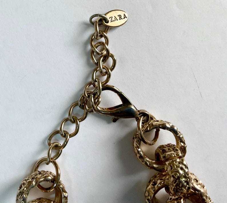Puede incluir: Primer plano de un collar dorado con cierre de mosquet&oacute;n y eslabones de cadena. El collar presenta un dise&ntilde;o texturizado y una peque&ntilde;a etiqueta con el nombre de la marca "ZARA". La joyer&iacute;a es probablemente para fines de moda o decoraci&oacute;n.