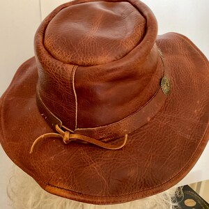 May include: A brown leather hat with a wide brim and a rounded crown. The hat has a decorative metal button and a leather tie. The leather has a textured, distressed appearance, suggesting a vintage or rustic style.