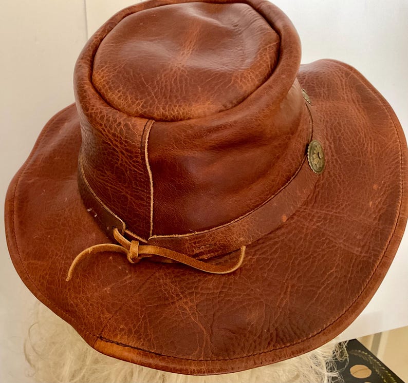 May include: A brown leather hat with a wide brim and a rounded crown. The hat has a decorative metal button and a leather tie. The leather has a textured, distressed appearance, suggesting a vintage or rustic style.