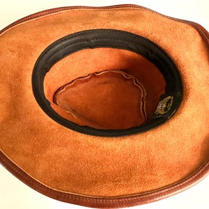 May include: A brown suede cowboy hat with dark brown leather trim and a black interior band. The hat has a classic western style, suitable for outdoor activities or fancy dress.