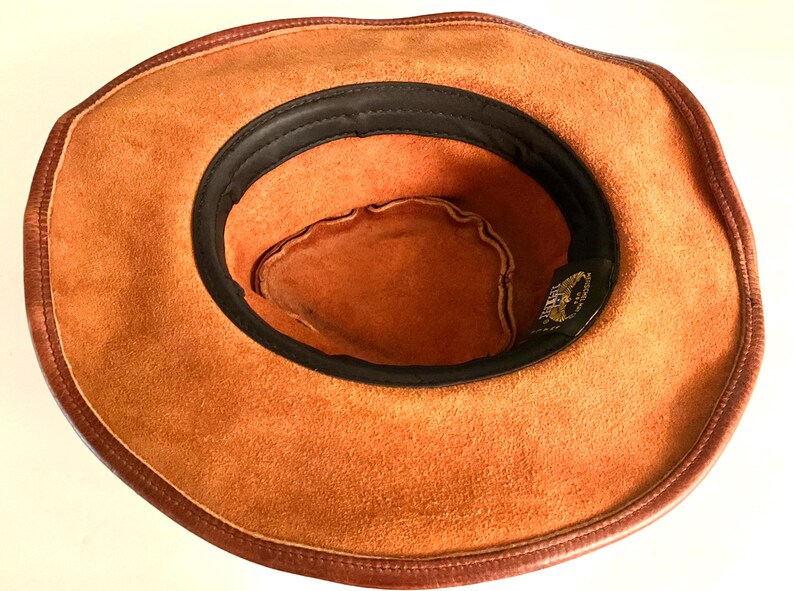 May include: A brown suede cowboy hat with dark brown leather trim and a black interior band. The hat has a classic western style, suitable for outdoor activities or fancy dress.