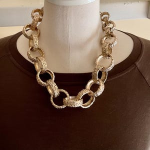 Puede incluir: Un collar dorado grueso con eslabones texturizados se muestra en un maniqu&iacute;. El collar est&aacute; compuesto por grandes anillos interconectados, creando un dise&ntilde;o audaz. El maniqu&iacute; lleva una camiseta marr&oacute;n de manga larga.