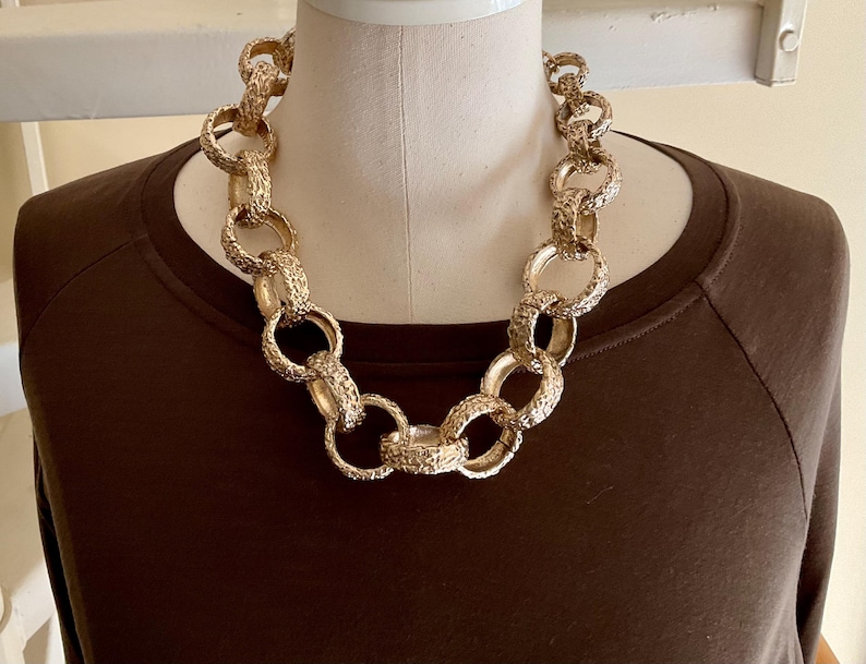 Puede incluir: Un collar dorado grueso con eslabones texturizados se muestra en un maniqu&iacute;. El collar est&aacute; compuesto por grandes anillos interconectados, creando un dise&ntilde;o audaz. El maniqu&iacute; lleva una camiseta marr&oacute;n de manga larga.