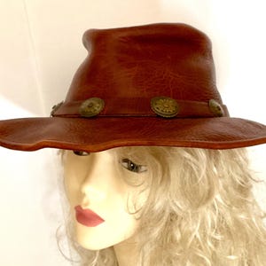 May include: A brown leather hat with a wide brim and a slightly curved crown. The hat features decorative metal buttons around the band. The hat has a handmade appearance and a rustic, vintage aesthetic.