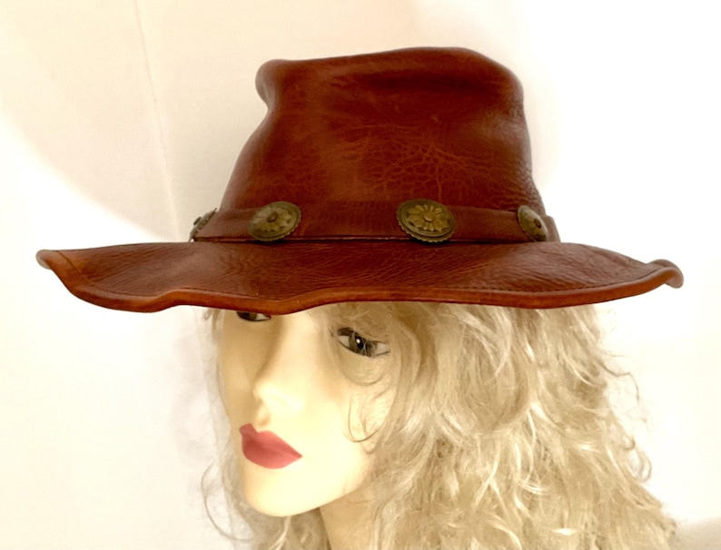 May include: A brown leather hat with a wide brim and a slightly curved crown. The hat features decorative metal buttons around the band. The hat has a handmade appearance and a rustic, vintage aesthetic.