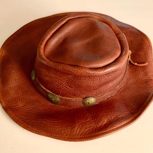 May include: A brown leather cowboy hat with a wide brim and a rounded crown. The hat has a decorative band with three brass-coloured studs. The leather has a textured appearance and a natural, worn look.