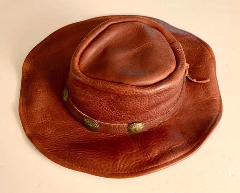 May include: A brown leather cowboy hat with a wide brim and a rounded crown. The hat has a decorative band with three brass-coloured studs. The leather has a textured appearance and a natural, worn look.