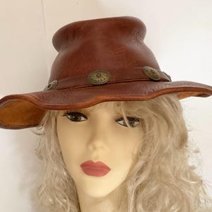 May include: A brown leather hat with a wide brim and a rounded crown. The hat features decorative metal accents around the brim. The hat is a western style and is likely handmade.