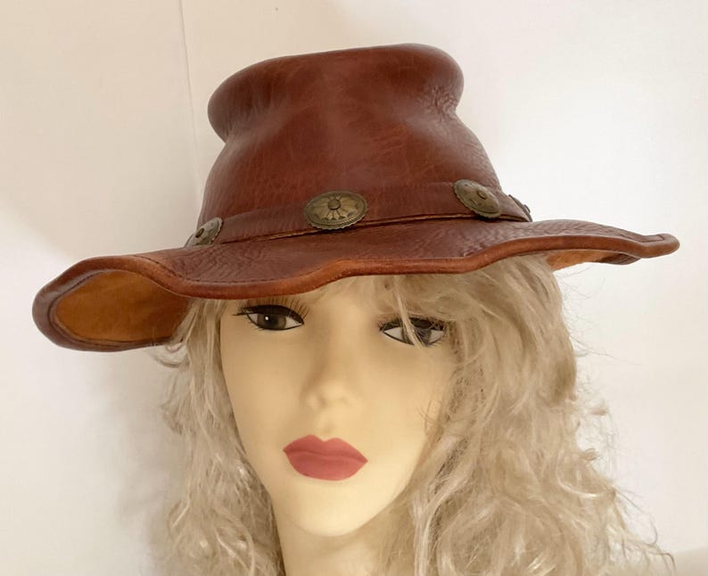 May include: A brown leather hat with a wide brim and a rounded crown. The hat features decorative metal accents around the brim. The hat is a western style and is likely handmade.