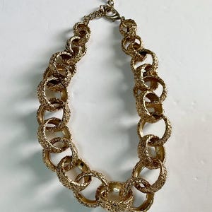 Puede incluir: Un collar dorado con grandes eslabones ovalados texturizados e interconectados. El collar tiene un cierre de mosquet&oacute;n y una cadena ajustable. La joya se exhibe sobre una superficie blanca.