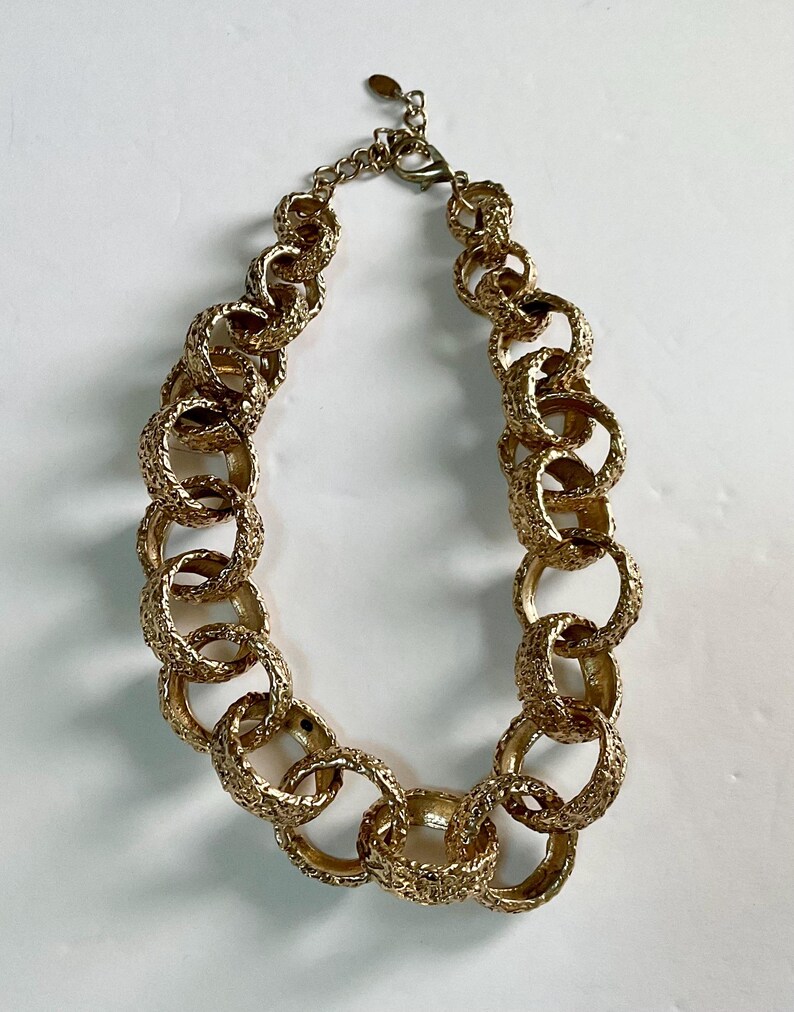 Puede incluir: Un collar dorado con grandes eslabones ovalados texturizados e interconectados. El collar tiene un cierre de mosquet&oacute;n y una cadena ajustable. La joya se exhibe sobre una superficie blanca.