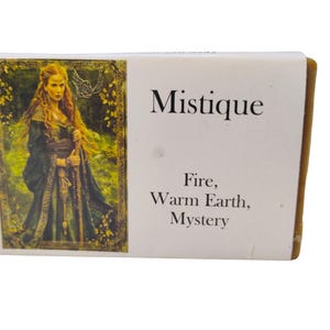 May include: A rectangular soap bar with a green and gold label featuring a woman in a green dress and the text "Mystique". The label also includes the text "Fire, Warm Earth, Mystery".