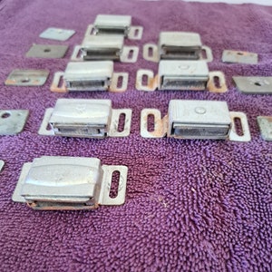 May include: A close-up of several silver magnetic cabinet door catches. The catches are arranged in rows on a purple textured surface.