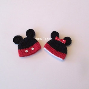 May include: Two crocheted Mickey Mouse hats, one black with white buttons and one black with a red bow and white polka dots. The hats are on a white background.