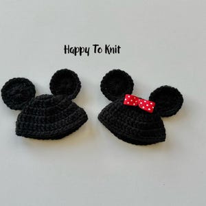 May include: Two black crocheted hats with mouse ears. One hat has a red and white polka dot bow. The hats are handmade and are likely for a baby or child. The text "Happy To Knit" is above the hats.