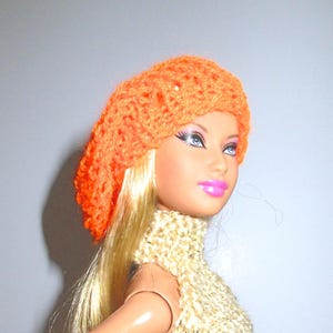 May include: A doll wearing a bright orange crocheted beret and a cream crocheted scarf.
