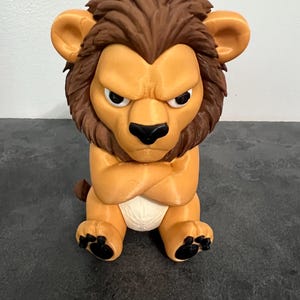 May include: A 3D printed figurine of a seated lion with a grumpy facial expression, brown mane, tan body, and a white diaper. The lion has its arms crossed over its chest, black nose, and black paw pads on its feet, rendered in a cartoon style.