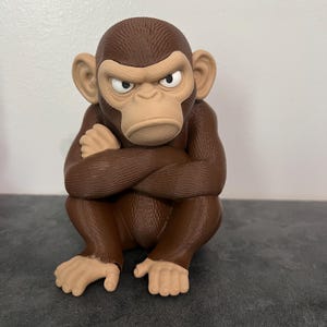 May include: A brown monkey figurine with a textured surface, depicted in a seated position with arms crossed over its chest. The monkey has a light tan face, ears, and hands, with a stern facial expression featuring narrowed eyes and a downturned mouth.