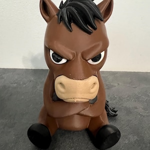 May include: A brown horse figurine with a black mane and tail, featuring a grumpy facial expression with furrowed brows and crossed arms. The horse has a light tan muzzle, white eyes with black pupils, and black hooves, sitting in a seated position.