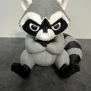 May include: A 3D printed raccoon figurine in shades of gray, black, and white. The raccoon is depicted in a seated position with its arms crossed over its chest, featuring a textured fur pattern, black eye patches, and a striped tail.