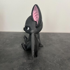 K&ouml;nnte beinhalten: Eine kleine, schwarze, stilisierte Fledermausfigur in Seitenansicht. Die Figur hat einen strukturierten K&ouml;rper, gro&szlig;e Ohren mit hellrosa Innendetails und gefaltete Fl&uuml;gel. Die Fledermaus hat einen runden Kopf, eine kleine Nase und ein sichtbares Auge.