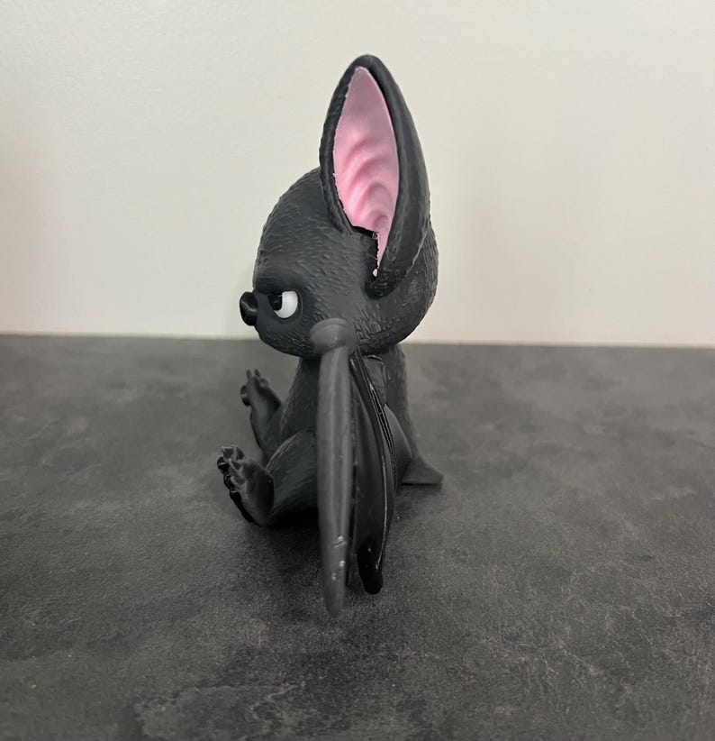 K&ouml;nnte beinhalten: Eine kleine, schwarze, stilisierte Fledermausfigur in Seitenansicht. Die Figur hat einen strukturierten K&ouml;rper, gro&szlig;e Ohren mit hellrosa Innendetails und gefaltete Fl&uuml;gel. Die Fledermaus hat einen runden Kopf, eine kleine Nase und ein sichtbares Auge.