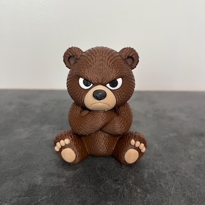 May include: A small brown bear figurine with a textured fur finish, depicted in a seated position with arms crossed over its chest. The bear has a stern facial expression with furrowed brows, white eyes, and a light tan muzzle and paw pads.