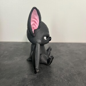 K&ouml;nnte beinhalten: Eine kleine schwarze Fledermausfigur mit strukturiertem Fell, gro&szlig;en spitzen Ohren mit rosa Innendetails und wei&szlig;en Augen. Die Fledermaus ist in sitzender Position dargestellt, wobei die Fl&uuml;gel am K&ouml;rper anliegen, in einer Seitenansicht.