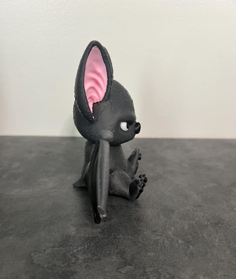 K&ouml;nnte beinhalten: Eine kleine schwarze Fledermausfigur mit strukturiertem Fell, gro&szlig;en spitzen Ohren mit rosa Innendetails und wei&szlig;en Augen. Die Fledermaus ist in sitzender Position dargestellt, wobei die Fl&uuml;gel am K&ouml;rper anliegen, in einer Seitenansicht.