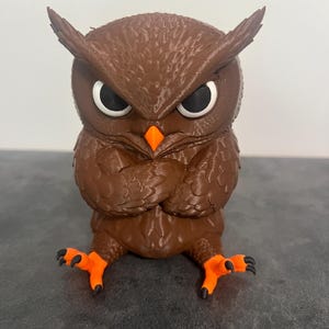 May include: A brown owl figurine with a textured feather pattern, large white eyes with black pupils, a bright orange beak, and orange feet with black talons. The owl is depicted with its wings folded across its chest in a seated position.