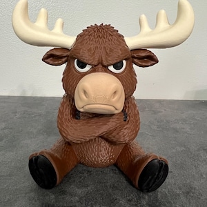 May include: A brown moose figurine with large cream-colored antlers, a textured fur-like surface, and a grumpy facial expression. The moose is depicted in a seated position with its arms crossed over its chest and black hooves.