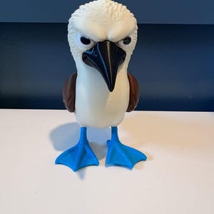 May include: A figurine of a blue-footed booby bird with a textured white head, a sharp black beak, brown wings, and bright blue webbed feet. The bird has a stern facial expression with narrowed eyes, standing upright on a flat surface.