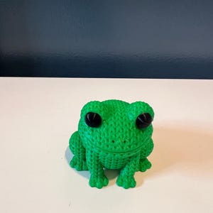 May include: A green frog figurine with a knitted texture and large black eyes. The frog is sitting on a white surface, with a dark blue background. The frog is a decorative item.
