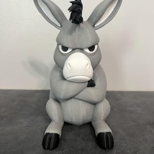 May include: A gray donkey figurine with a white muzzle, black hooves, and a black tuft of hair on its head. The donkey has a stern facial expression with furrowed brows and its arms are crossed over its chest in a seated, upright position.
