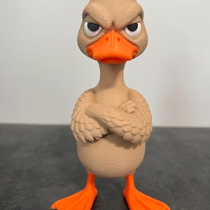 May include: A tan-colored duck figurine with a textured surface, featuring a small red crest on its head, bright orange webbed feet, and an orange beak. The duck has a stern facial expression with furrowed brows and its wings are crossed.