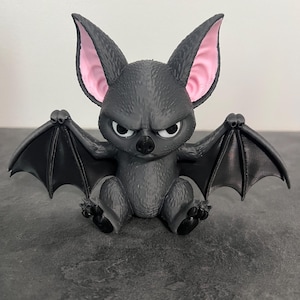 May include: A small, dark gray bat figurine with a textured, fur-like body and large, pointed ears featuring light pink inner coloring. The bat has a stern facial expression with white eyes, a black nose, and outstretched wings with visible webbing.
