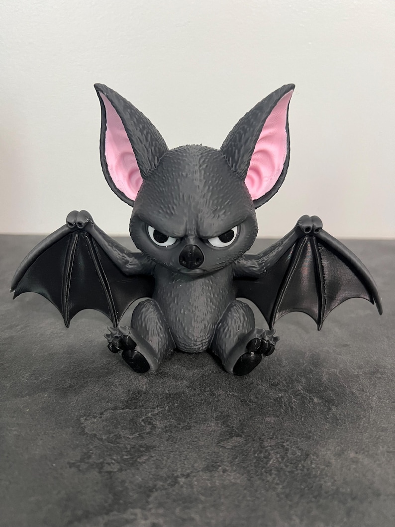 K&ouml;nnte beinhalten: Eine kleine, dunkelgraue Fledermausfigur mit strukturiertem, fellartigem K&ouml;rper und gro&szlig;en, spitzen Ohren mit hellrosa Innenseite. Die Fledermaus hat einen strengen Gesichtsausdruck mit wei&szlig;en Augen, einer schwarzen Nase und ausgebreiteten Fl&uuml;geln.