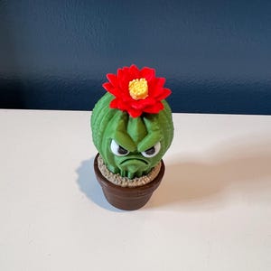 May include: A green cactus figurine with a textured surface and a grumpy facial expression, featuring white eyes with black pupils and a downturned mouth. A bright red flower with a yellow center sits on top, all housed in a brown pot.