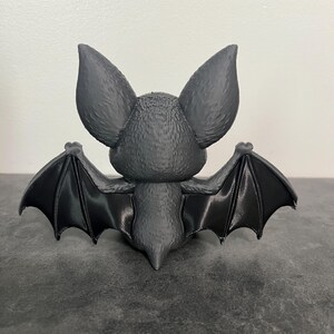 K&ouml;nnte beinhalten: Eine mattschwarze Fledermausfigur, von hinten betrachtet. Die Fledermaus hat gro&szlig;e, texturierte Ohren, einen kleinen K&ouml;rper mit feinen fellartigen Details und vollst&auml;ndig ausgebreitete Fl&uuml;gel mit sichtbarer Strukturierung auf der Oberfl&auml;che.