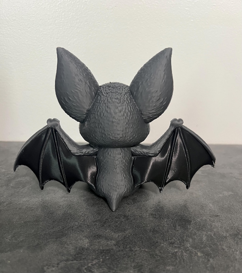 K&ouml;nnte beinhalten: Eine mattschwarze Fledermausfigur, von hinten betrachtet. Die Fledermaus hat gro&szlig;e, texturierte Ohren, einen kleinen K&ouml;rper mit feinen fellartigen Details und vollst&auml;ndig ausgebreitete Fl&uuml;gel mit sichtbarer Strukturierung auf der Oberfl&auml;che.