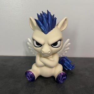 May include: A small, cream-colored figurine of a winged creature with a grumpy expression, arms crossed, and a metallic blue mane and tail. The figure features white wings, pointed ears, and metallic purple hooves, sitting in a seated position.
