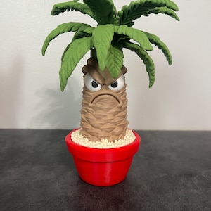 May include: A small figurine of a palm tree with a textured brown trunk, a grumpy facial expression with white eyes and a downturned mouth, and green fronds. The tree is planted in a bright red pot filled with small white pebbles.
