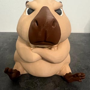 May include: A light tan capybara figurine with a dark brown nose and feet. The figure is depicted in a seated position with its arms crossed over its chest, featuring a stern facial expression with dark, focused eyes and a prominent snout.