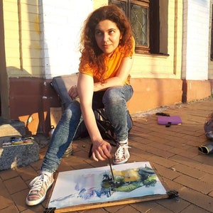 May include: A woman sits on a folding chair outside, painting a watercolor landscape on a canvas. She is wearing a bright orange shirt and blue jeans.