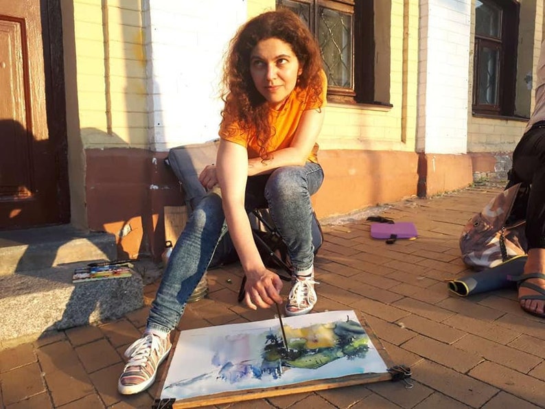 May include: A woman sits on a folding chair outside, painting a watercolor landscape on a canvas. She is wearing a bright orange shirt and blue jeans.