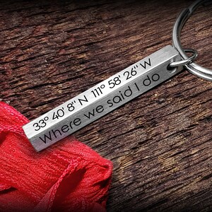 May include: A silver rectangular keychain engraved with the text "33° 40' 8" N 111° 58' 26" W Where we said I do." The keychain is attached to a silver ring. The background is a dark wooden surface with a red fabric.