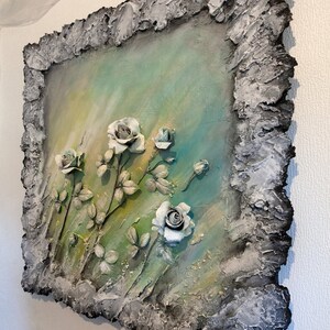 May include: A textured, gray frame surrounds a painting of white roses on a green and blue background. The roses are sculpted and appear to be three-dimensional.