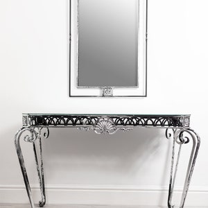 Könnte beinhalten: Ein dekoratives Konsolentisch- und Spiegel-Set aus Metall. Der Tisch hat eine Glasplatte und verzierte Beine. Der rechteckige Spiegel hat dekorative Details. Das Set ist in einem silbernen Farbton gehalten.
